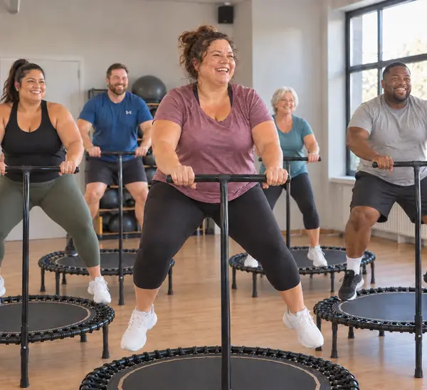 Lächelnde Frau springt entspannt auf einem Mini-Trampolin mit Haltestange, umgeben von einer diversen Gruppe – Stimmungsbild für den Kurs Curvy Jumping